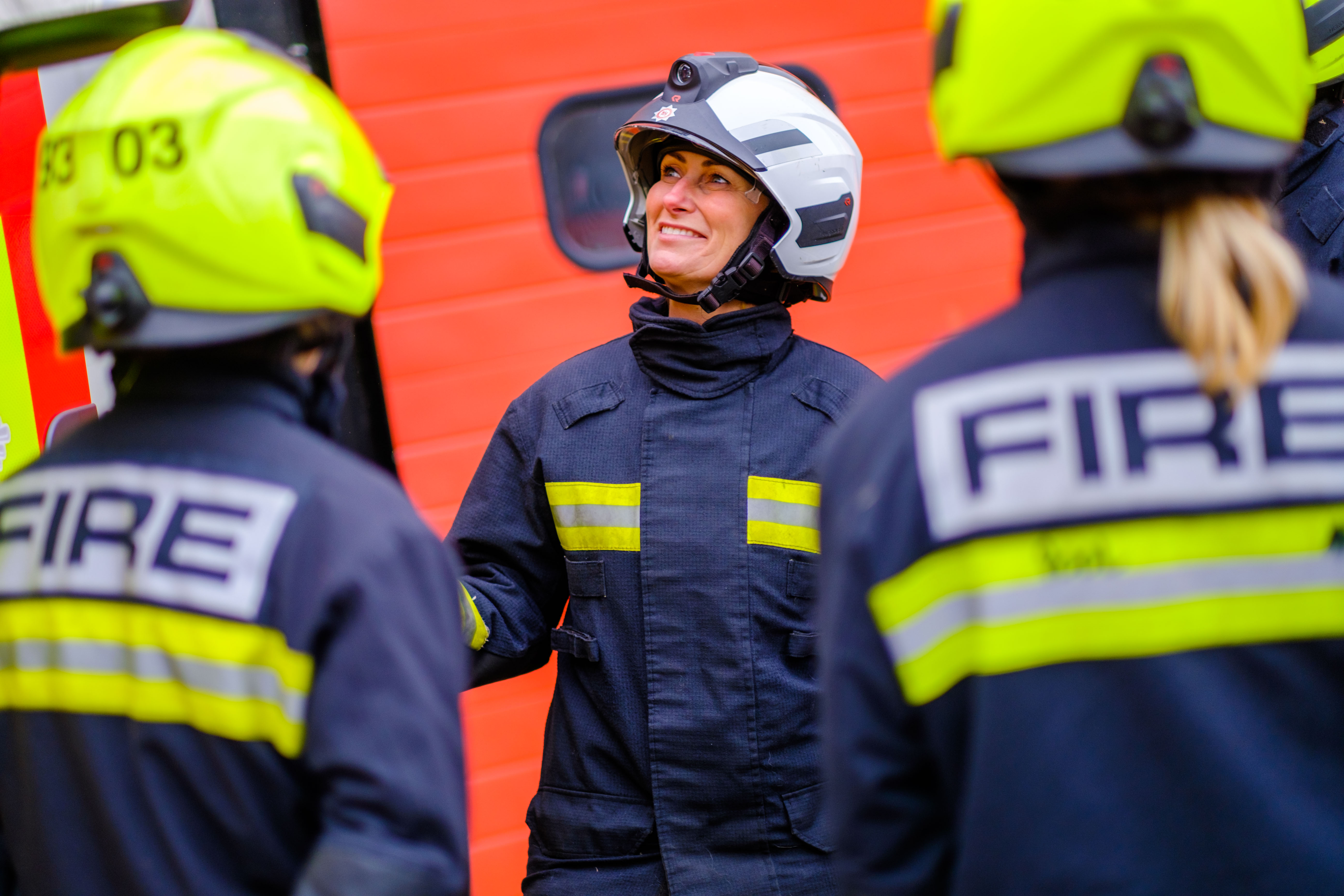 Three firefighters are pictured. A female firefighter with a white helmet is shown in the centre of the photo. She is speaking to two firefighters who are positioned on each side of her showing their 'Fire' jackets to the camera. 