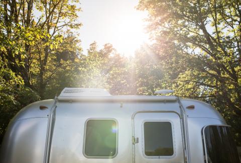 A silver mobile home underneath green trees and sunlight.