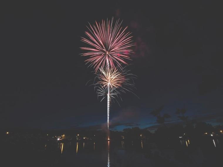 A red firework in the night sky, with orange and yellow smaller fireworks directly beneath it. The display looks like it's happening near a body of water.