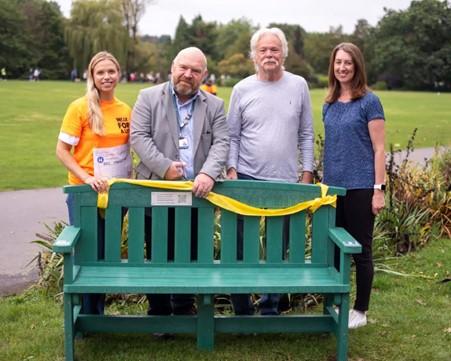 Four people standing by a green bench in a park