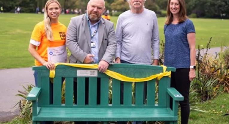 Four people standing by a green bench in a park